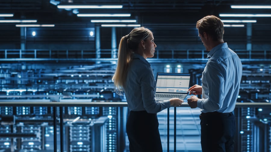 A couple of IT Techs monitoring data analytics while standing over a room filled as a server farm